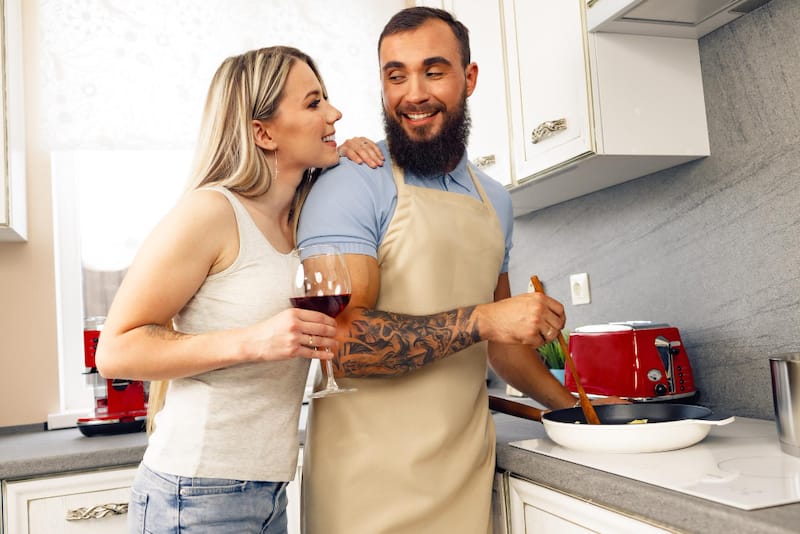 A young couple is in the kitchen, happy and enjoying each other's company as the husband cooks a meal.